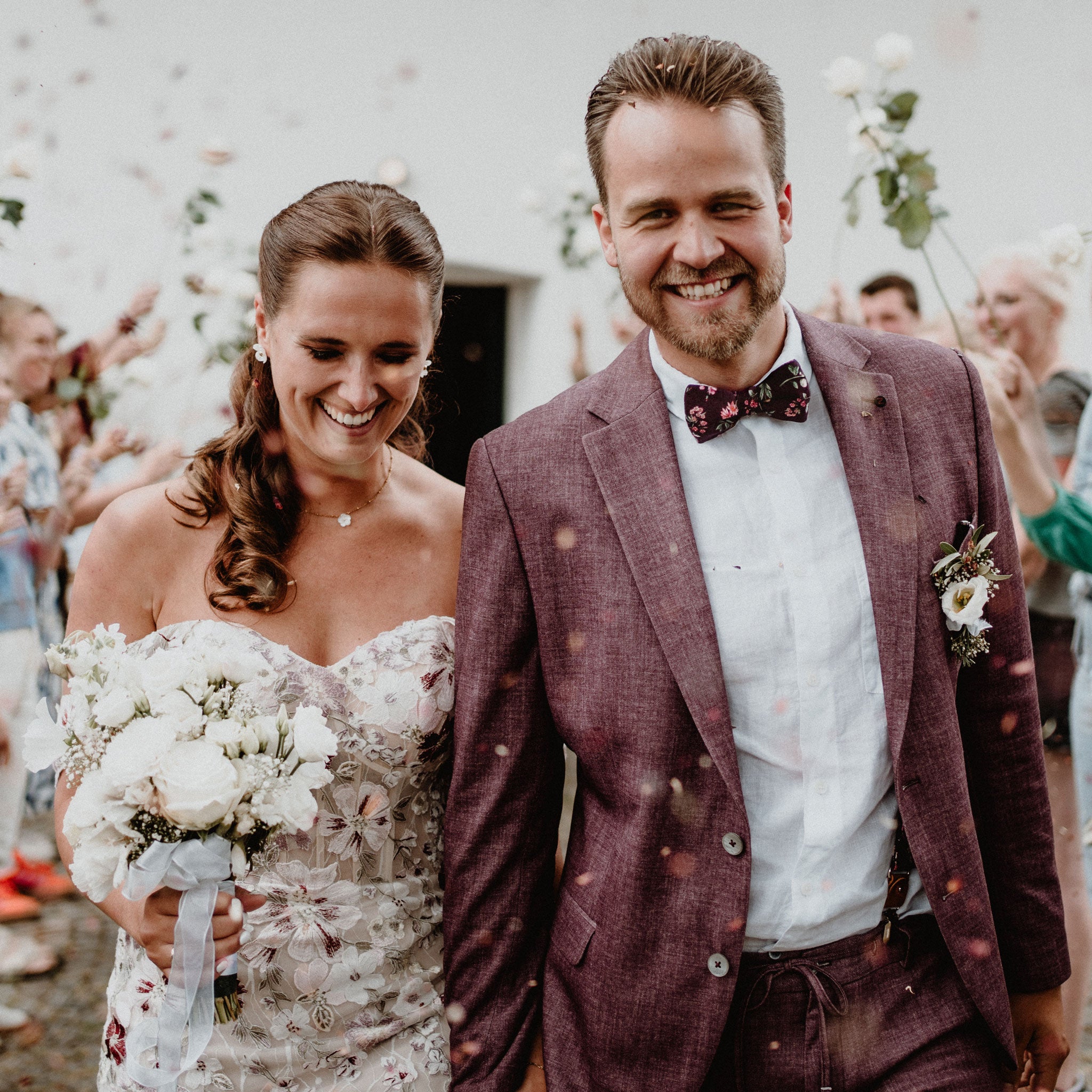 Brautpaar zwischen Hochzeitsgästen - Bräutigam mit blumiger fliege passend zum Anzug und Braut mit Hochzeitskleid